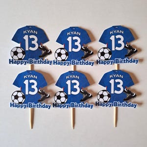 May include: Six blue soccer jersey cupcake toppers with the name "KYAN" and the number "13". Each topper includes a soccer ball, cleats, and the text "Happy Birthday". The toppers are on wooden sticks.