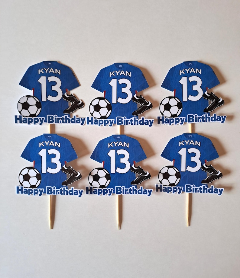 May include: Six blue soccer jersey cupcake toppers with the name "KYAN" and the number "13". Each topper includes a soccer ball, cleats, and the text "Happy Birthday". The toppers are on wooden sticks.