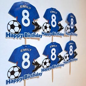 May include: Blue soccer-themed birthday cake toppers featuring a jersey with the name "EMILY" and the number "8", soccer balls, and cleats. The toppers also include the text "Happy Birthday".