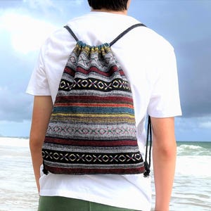 May include: A drawstring backpack featuring a woven fabric with horizontal stripes in black, white, red, blue, and yellow. The bag has a repeating geometric pattern of small crosses and lines, with black cord straps that cinch the top opening closed.