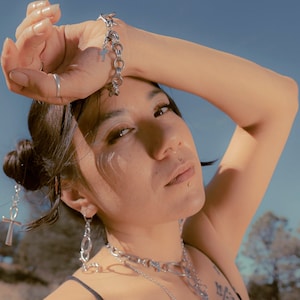 May include: A person wearing a black tank top with multiple silver chain necklaces and earrings. The person is wearing a silver chain bracelet on their wrist and has their arm raised above their head. The person is standing in a field of tall grass with trees in the background.