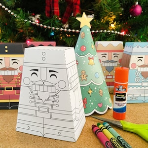 Pode incluir: Um conjunto de cinco caixas de artesanato de papel com tema natalino com designs de quebra-nozes e árvores de Natal. As caixas estão prontas para serem coloridas e montadas. A imagem também mostra cola, tesoura e lápis de cor.