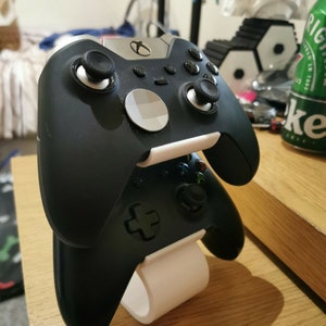 May include: Two black Xbox controllers are displayed on a white stand. The controllers are stacked on top of each other, with the top controller facing forward and the bottom controller facing backward. The stand is made of plastic and has a circular base.