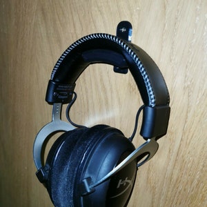 May include: Black and silver gaming headset with a black headband and earcups. The headset is hanging on a black metal hook attached to a wooden wall.