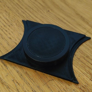 Puede incluir: Un objeto negro impreso en 3D con una parte superior circular y una base redondeada. El objeto está diseñado para sostener un objeto pequeño, como un anillo o una moneda.