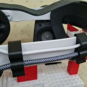 Puede incluir: Primer plano de un auricular VR blanco con correas negras y un cable negro. El cable está asegurado al auricular con dos clips de cable negros.