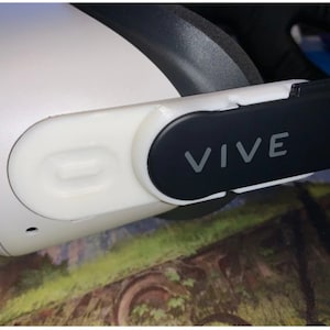 May include: A white VR headset with a black strap and a black cover with the word "VIVE" printed on it.