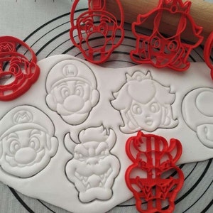 Puede incluir: Conjunto de nueve cortadores de galletas de plástico rojo y blanco con personajes de la franquicia de videojuegos Super Mario Bros. Los cortadores incluyen a Mario, Luigi, la princesa Peach, Toad, Bowser y un bloque de interrogación.