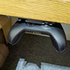 Puede incluir: Un mando de videojuegos negro está montado debajo de un escritorio de madera con un clip. El mando está colgado boca abajo con los botones mirando hacia el techo.