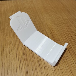May include: White plastic stand with a PlayStation logo on the front. The stand is designed to hold a video game controller.