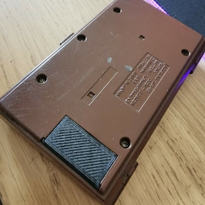 Könnte beinhalten: Ein braunes, handliches elektronisches Spiel mit einem schwarzen Gummiknopf an der Seite. Das Spiel hat Text auf der Rückseite, der "NPEEP NI SORIN", "EXPRESS PRESS ONE", "MODEL NO.: JR-35", "ABELIVE", "SATA" und "TING" lautet.
