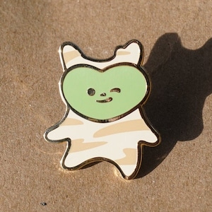 Op de afbeelding: Een gouden emaille pin met een cartoon dier met een hartvormig hoofd. Het dier draagt een gestreept patroon en heeft een knipperend oog. Het hart is groen.