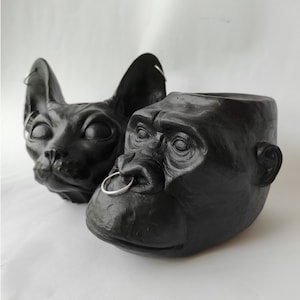 May include: Two black decorative planters, one shaped like a cat's head and the other a gorilla's head. Both have a silver ring in their nose. The cat planter has large ears and the gorilla planter has detailed facial features.