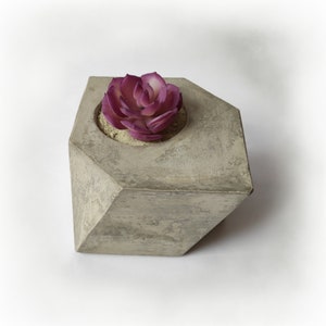 May include: A geometric concrete planter with a faux succulent. The planter is a light gray color with a textured finish. The succulent has deep purple and white petals, and sits in a bed of sand.