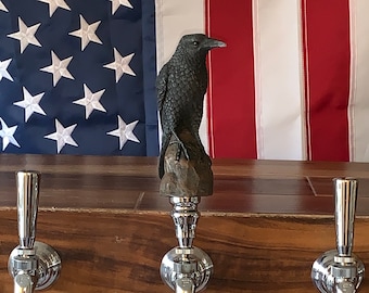 May include: A dark, detailed raven sculpture atop a beer tap handle. The tap is made of polished metal with a black handle. The background features an American flag and a wooden bar.