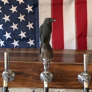 May include: A dark, detailed raven sculpture atop a beer tap handle. The tap is made of polished metal with a black handle. The background features an American flag and a wooden bar.