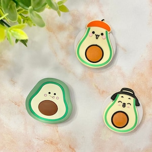 May include: Three avocado-shaped accessories with smiling faces. One avocado wears a beret, another a hat, and the third has no accessories. The avocados are green with brown centres and a white background.