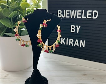 May include: Gold hoop earrings with red, green, and white beads. The earrings are on a black display stand. The text "BEJEWELED BY KIRAN" is on a black letter board behind the earrings.
