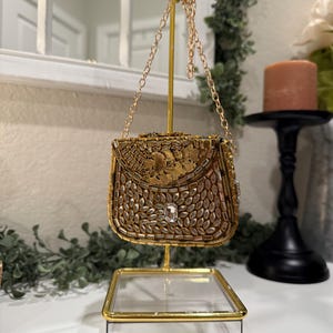 May include: A small, ornate gold handbag with a chain strap, displayed on a gold stand. The bag is embellished with intricate beadwork and a central jewel. The stand has a clear acrylic base with gold trim, showcasing the bag's details.