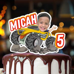 May include: A cake topper featuring a cartoon image of a child in a yellow monster truck with the name "MICAH" and the number "5". The cake has white frosting with chocolate ganache dripping down the sides.