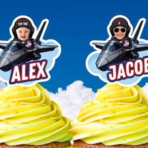 Puede incluir: Dos cupcakes con glaseado amarillo, cada uno coronado con una imagen de dibujos animados de un niño en un avión de combate. Una imagen tiene el nombre "ALEX" y la otra "JACOB" en texto rojo y blanco. El fondo es un cielo azul con nubes.