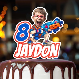 May include: A cake topper featuring a cartoon illustration of a boy holding a toy blaster. The number "8" and the name "JAYDON" are prominently displayed. The cake is topped with chocolate ganache.