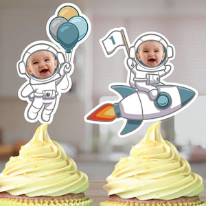 May include: Two cupcake toppers featuring an astronaut with a rocket and an astronaut holding balloons. The astronaut's faces are blank and can be personalized with photos. The toppers are perfect for a first birthday party.