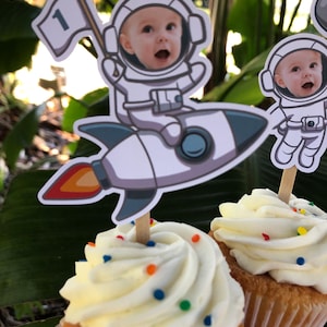 May include: Two cupcakes with white frosting and colorful sprinkles topped with astronaut-themed cake toppers. One topper features a baby in an astronaut suit riding a rocket with a flag that says "1". The other topper features a baby in an astronaut suit.