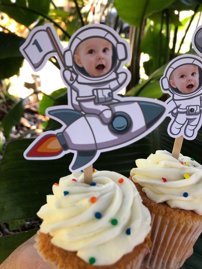 May include: Two cupcakes with white frosting and colorful sprinkles topped with astronaut-themed cake toppers. One topper features a baby in an astronaut suit riding a rocket with a flag that says "1". The other topper features a baby in an astronaut suit.