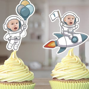 May include: Two astronaut cupcake toppers with a baby's face in the helmet. One astronaut is holding balloons and the other is riding a rocket with the number "1" on it.