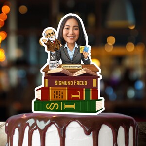 May include: A cake topper featuring a cartoon illustration of a woman in a blazer, standing on a stack of books labeled "Sigmund Freud". A smaller cartoon of a man is next to her. The cake is topped with chocolate frosting.