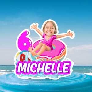 May include: A cake topper with a smiling girl in a pink swimsuit on a pink donut float. The number 6 and the name "MICHELLE" are displayed. The background shows a beach and blue sky.