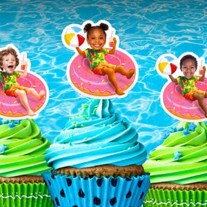 May include: Cupcakes with colorful frosting and decorative toppers featuring children in inflatable rings with beach balls. The cupcakes have green and blue frosting, and are set against a blue water background.