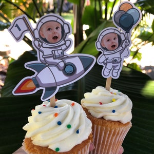 May include: Two white frosted cupcakes with colorful sprinkles, decorated with astronaut toppers. One astronaut is holding a flag with the number "1" on it, and the other is holding balloons.
