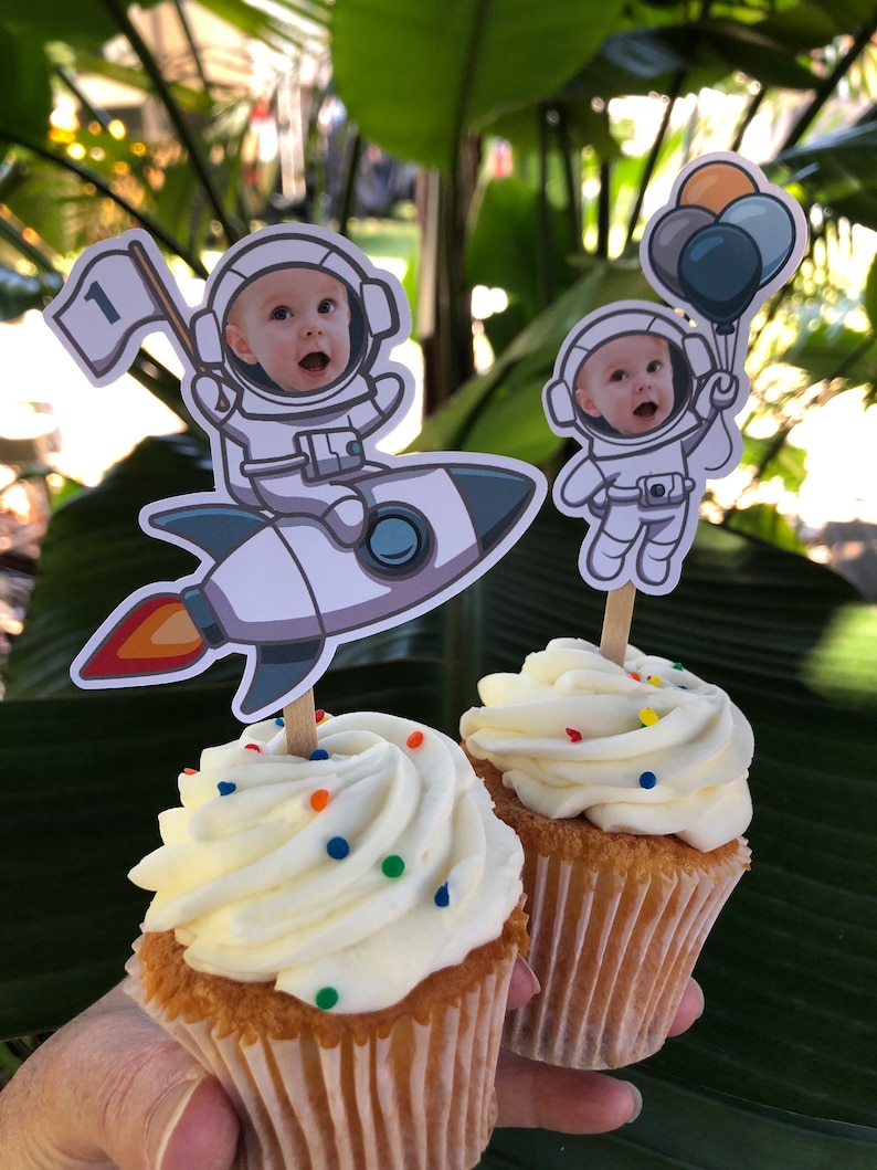 May include: Two white frosted cupcakes with colorful sprinkles, decorated with astronaut toppers. One astronaut is holding a flag with the number "1" on it, and the other is holding balloons.