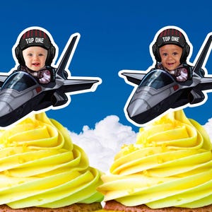 Puede incluir: Dos cupcakes cubiertos con glaseado amarillo. Cada uno tiene una imagen recortada de un bebé en un avión de combate, usando un casco con el texto "TOP ONE". El fondo es un cielo azul con nubes.