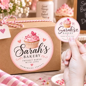 May include: A brown cardboard box featuring a large circular sticker with a pink cupcake illustration and the text Sarah's Bakery Freshly Baked With Love Homemade Treats. A smaller matching sticker is held up, showing the Sarah's Bakery logo.