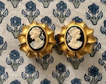 Könnte beinhalten: Zwei goldfarbene Cameo-Ohrringe. Jeder Ohrring hat ein ovales schwarzes Cameo mit einem weißen Profil einer Frau. Die Cameos sind in einem goldenen Rahmen mit einem Sonnenstrahl-Design gefasst. Die Ohrringe werden auf einem blau geblümten Stoff präsentiert.