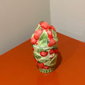 May include: A ceramic jar with a lid shaped like a bunch of strawberries. The jar is decorated with a green and white pattern of leaves and strawberries.