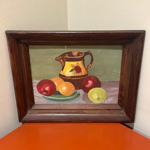 May include: A framed painting of a still life with a brown pitcher with yellow and red floral designs, three red apples, two yellow oranges, and a green apple on a green plate. The painting is signed "A. Guitter" in the bottom right corner.