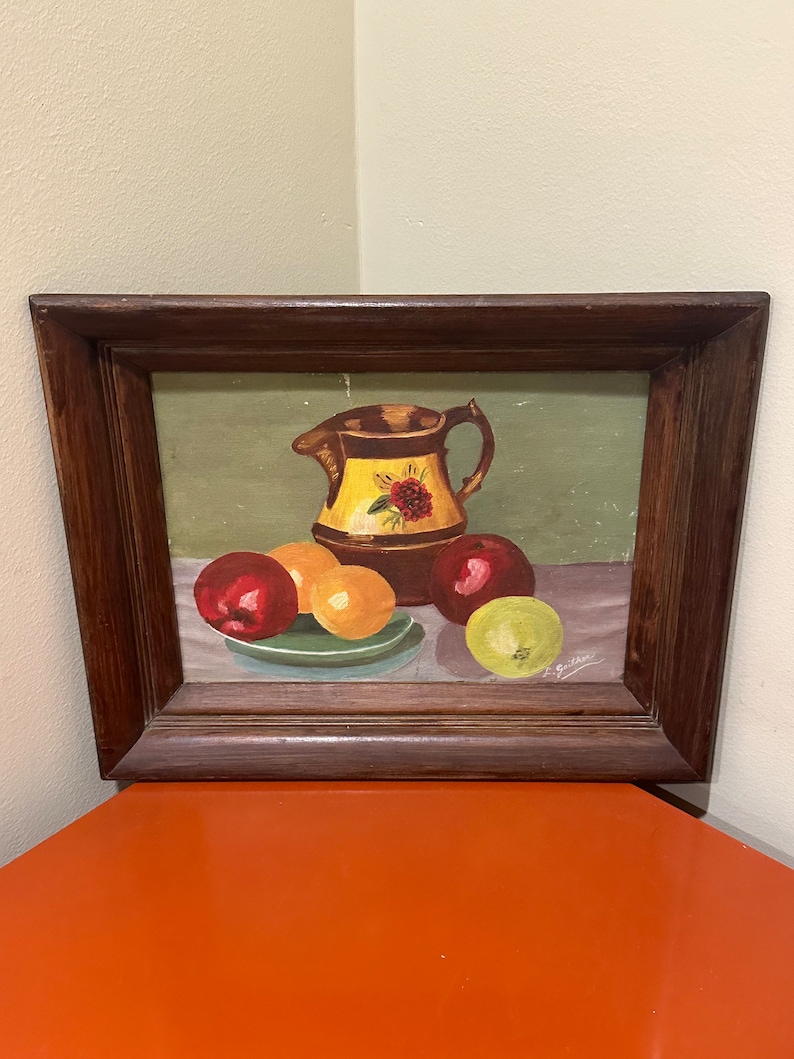 May include: A framed painting of a still life with a brown pitcher with yellow and red floral designs, three red apples, two yellow oranges, and a green apple on a green plate. The painting is signed "A. Guitter" in the bottom right corner.