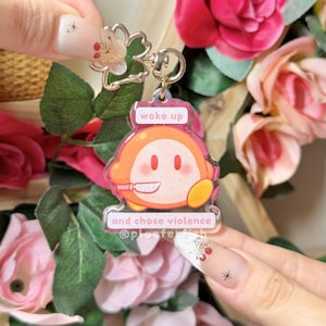 May include: A clear acrylic keychain featuring an orange cartoon character holding a knife. The keychain has the text "woke up" above the character and "and chose violence" below. The keychain is attached to a gold-colored metal clasp.