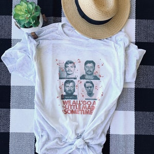 May include: A gray t-shirt with a graphic print of four mugshots with red blood splatter. The text "WE ALL GO A LITTLE MAD SOMETIME" is printed below the mugshots.