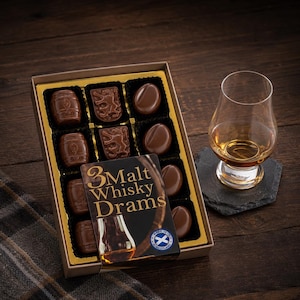 May include: A rectangular box of assorted milk chocolates featuring barrel and lion-shaped pieces, alongside a glass of amber liquid. The box lid displays the text 3 Malt Whisky Drams and a small circular logo with a blue and white cross.