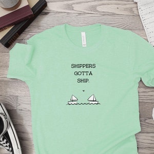 Può includere: Una maglietta verde chiaro con il testo "Shippers Gotta Ship." e un'immagine di due barche a vela.