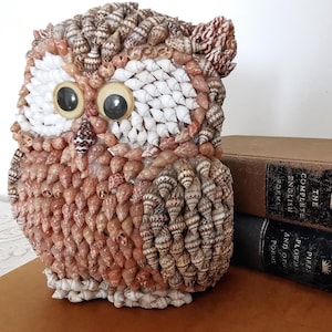 May include: A decorative owl figurine crafted from numerous small, layered seashells in shades of brown, tan, and white. The owl features two large, circular, smooth plastic eyes and a pointed beak made from a single dark, patterned seashell.