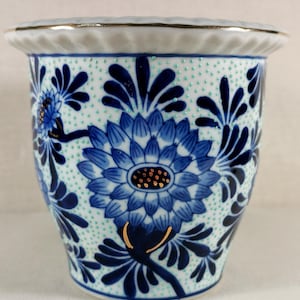 Op de afbeelding: Een keramische vaas met een witte basis, versierd met blauwe bloempatronen en gouden accenten. De vaas heeft een herhalend patroon van kleine groene stippen en een gouden rand. Het bloemmotief omvat een grote centrale bloem en kleinere omringende motieven.