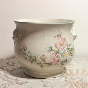 Puede incluir: Una maceta de cerámica color crema con cuerpo redondeado y borde ensanchado. El exterior presenta un delicado patrón floral de flores rosas y azules con hojas verdes. Dos pequeñas asas esculpidas se sitúan en los lados opuestos del recipiente.