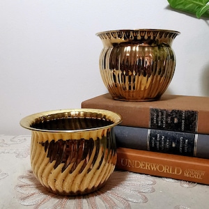 Può includere: Due vasi in ottone lucido con un design arrotondato e scanalato. Un vaso è posizionato sopra una pila di tre libri, mentre l'altro si trova davanti. I libri presentano dorsi con testo visibile, tra cui The Complete English Poems e Underworld.