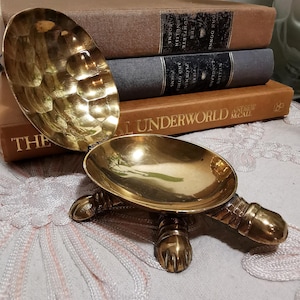 May include: A brass turtle-shaped trinket dish with a hinged shell that opens to reveal a smooth, polished interior. The exterior of the shell features a hammered texture, while the base includes four detailed legs and a head extending outward.